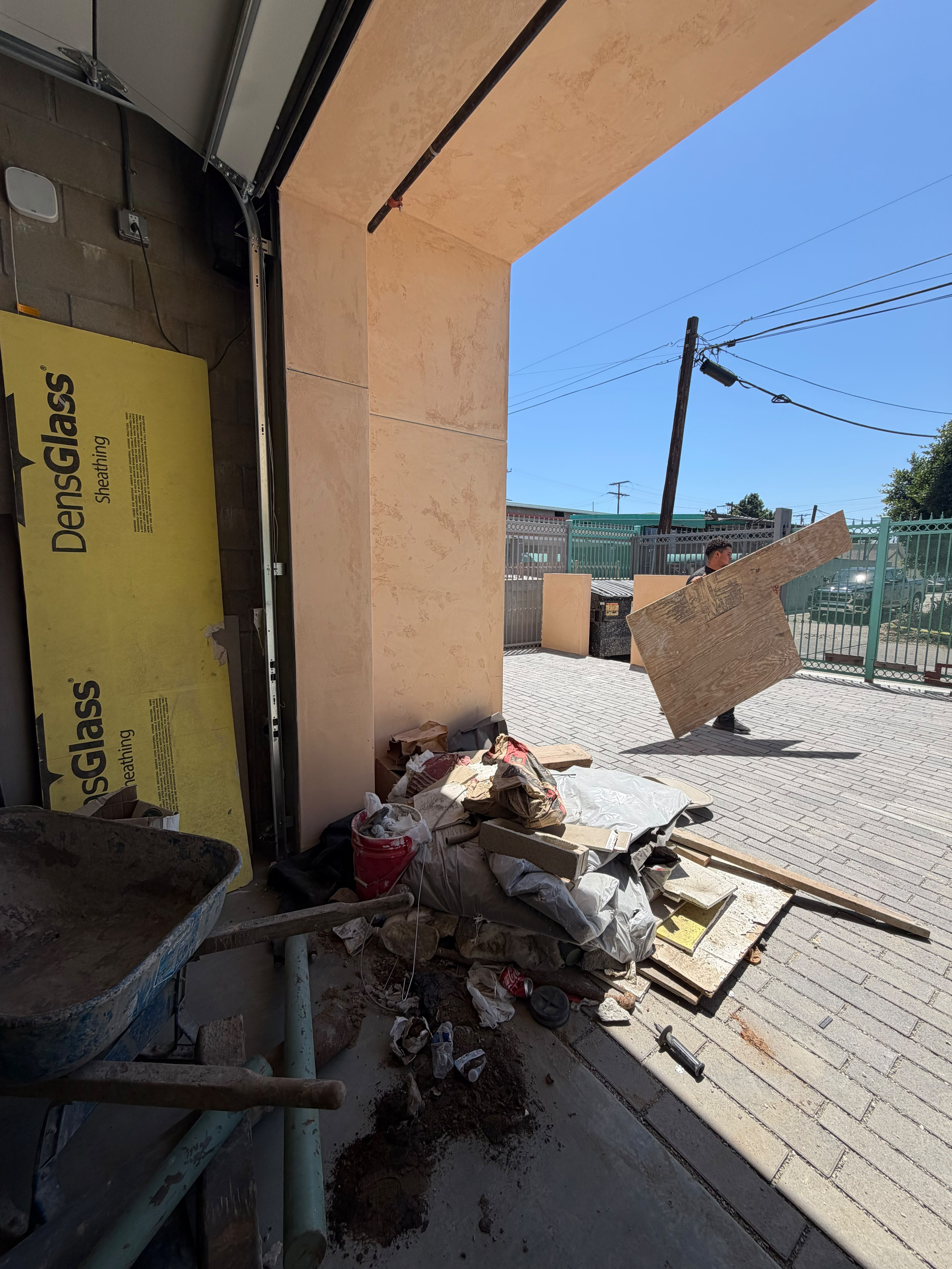 Worker carrying plywood past construction debris outside a garage doorway